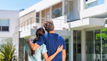 couple embracing in front of new big modern house, outdoor rear view back looking at their dream home